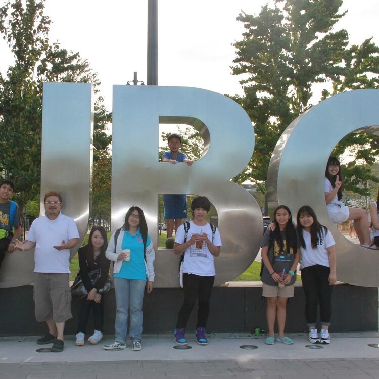 A group of PURE students, Ringo and Cheryl posing in front of a large UBC sign, representing a summer music institute partnership.