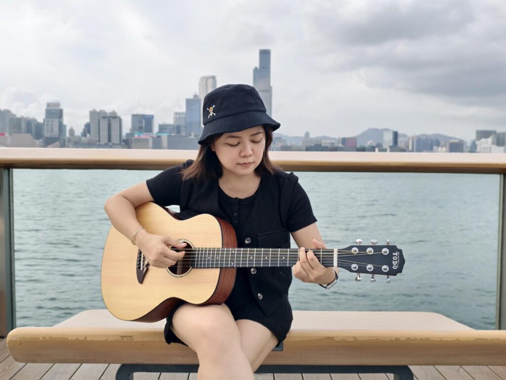 Lorraine is playing her guitar, sitting on the bench, back facing the sea.