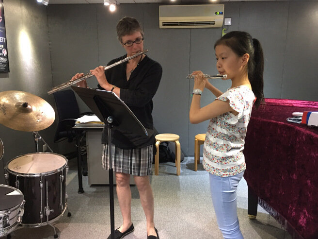 A female teacher giving a 1-on-1 music lesson on the flute to a young student in a music studio. Both are holding flutes and appearing to play.